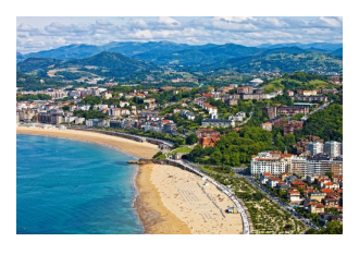 looking down onto a yellow beach, green hills, red roofed buildings against a blue sea and blue sky