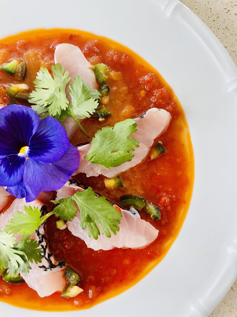 a white plate of food, brown orange sauce, white raw fish, green leaves and a purple flower
